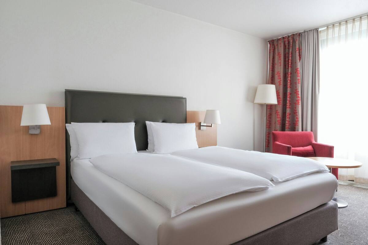A hotel room with a large bed, two bedside lamps, a red armchair, and a small table.