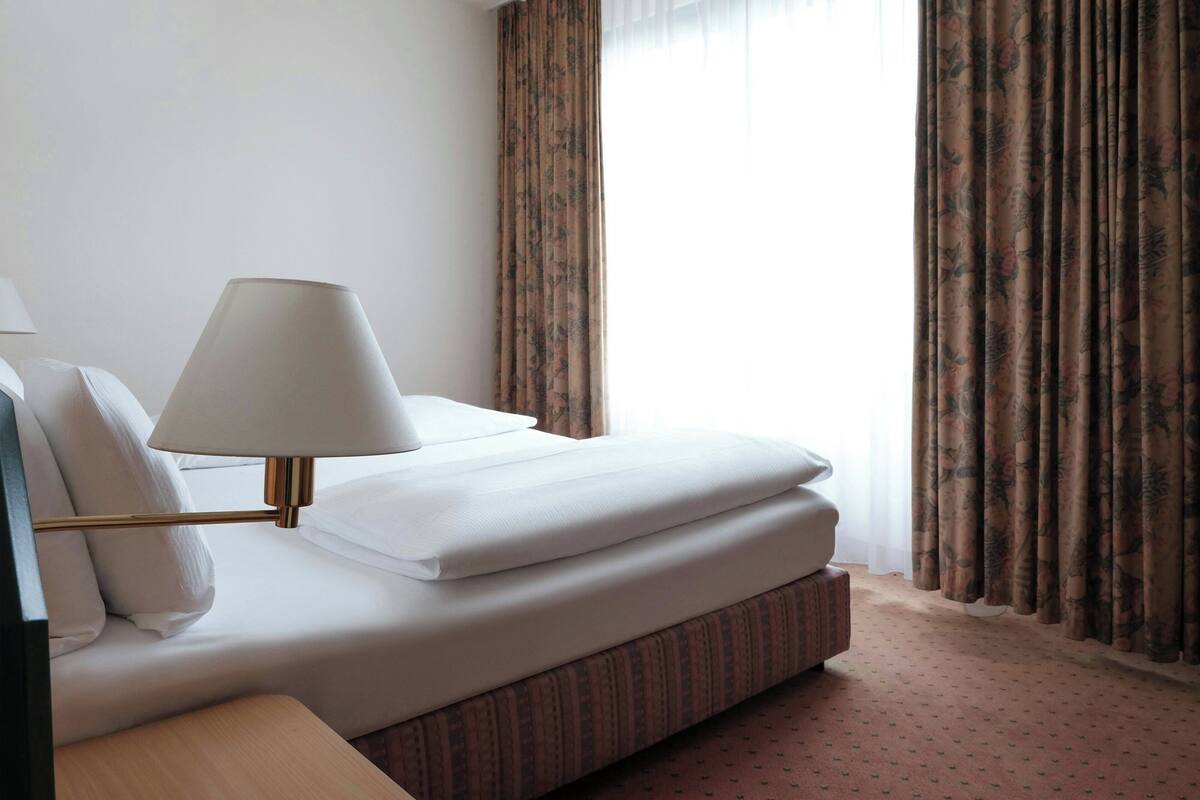 A neatly made bed with white linens, a bedside lamp, and a patterned curtain.