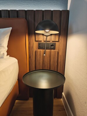 Una cama con una almohada blanca y una lámpara negra sobre una mesita de noche de madera.