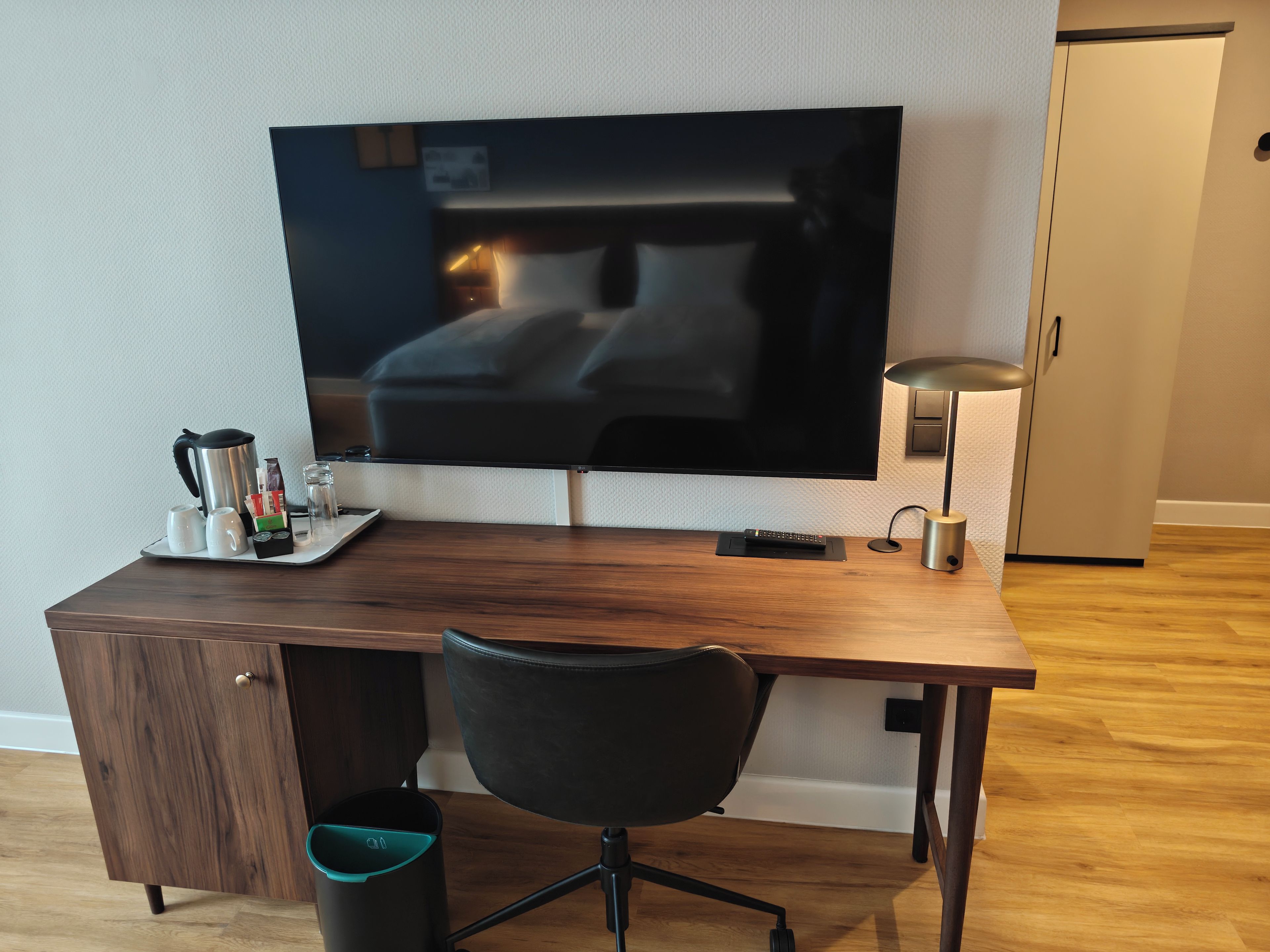 A hotel room with a wooden desk, a black office chair, a television, a coffee maker, and a trash bin.