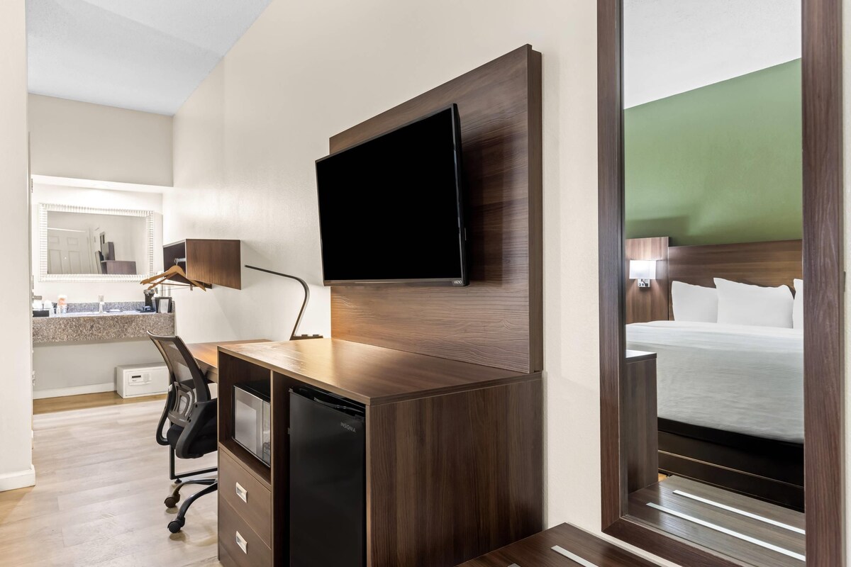 Una habitación de hotel con un escritorio de madera, un televisor de pantalla plana montado en la pared, una cama con ropa blanca y un espejo que refleja la habitación.