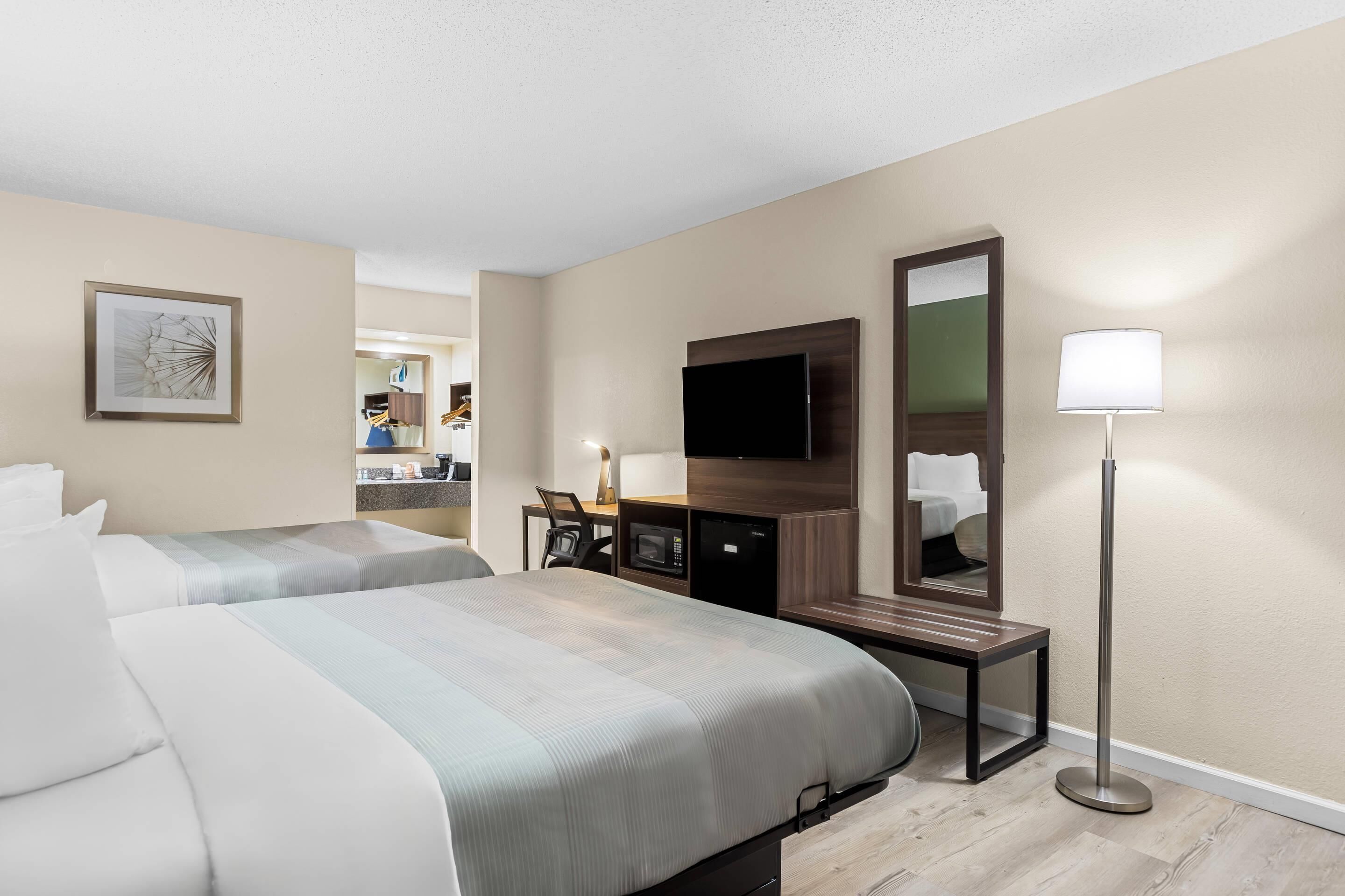A hotel room with two beds, a desk with a flat-screen TV, a mirror, and a floor lamp.