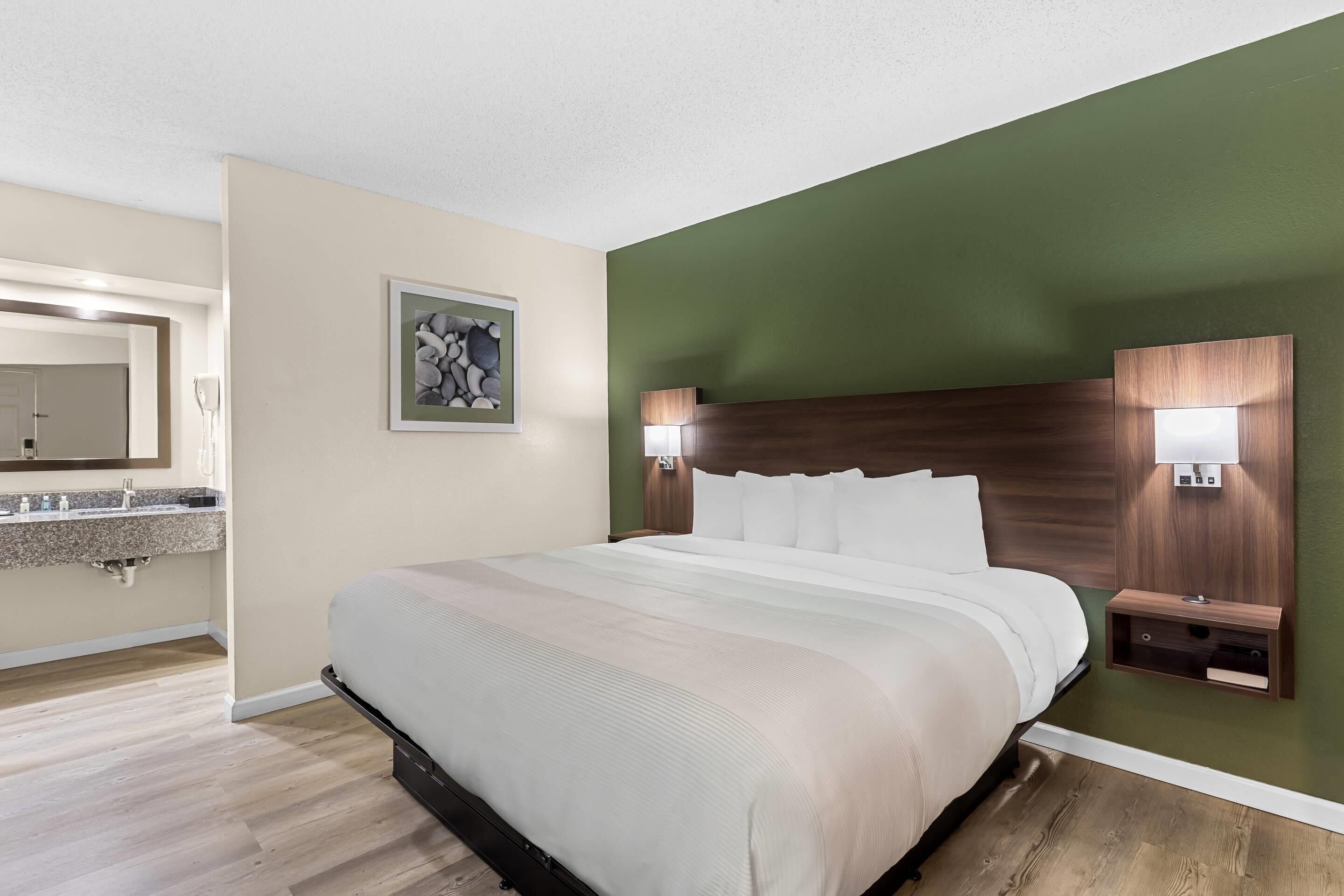 Een hotelkamer met een groot bed, een houten hoofdeinde, twee nachtkastjes met lampen, een schilderij aan de muur en een badkamer die door een open deur zichtbaar is.