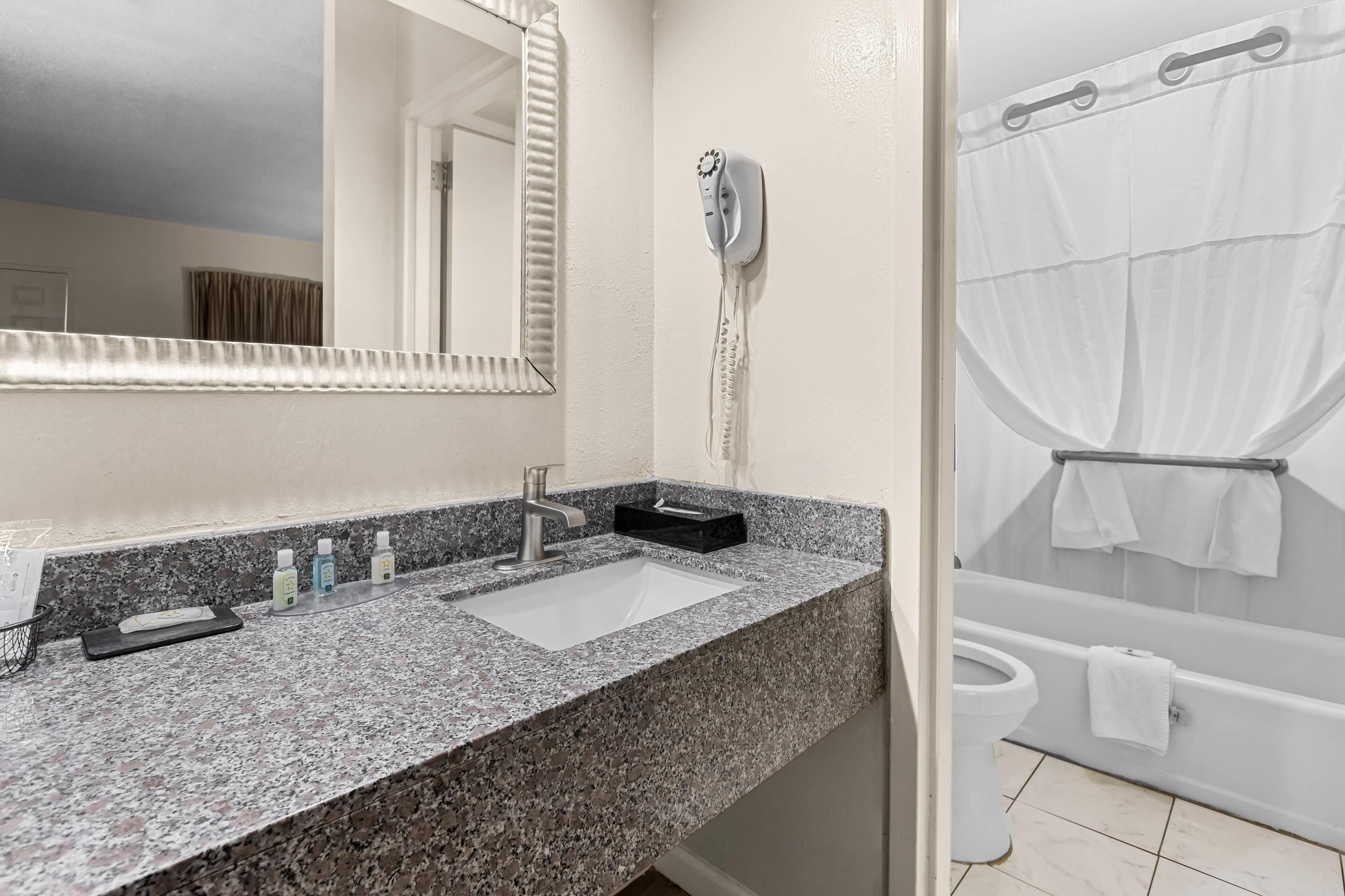 Un baño con encimera de granito, lavamanos, secador de pelo y bañera con cortina de ducha.