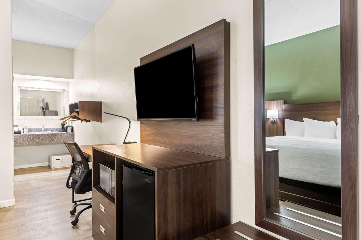 Habitación de hotel con escritorio de madera, televisor de pantalla plana montado en la pared, refrigerador negro pequeño y cama con cabecera de madera.