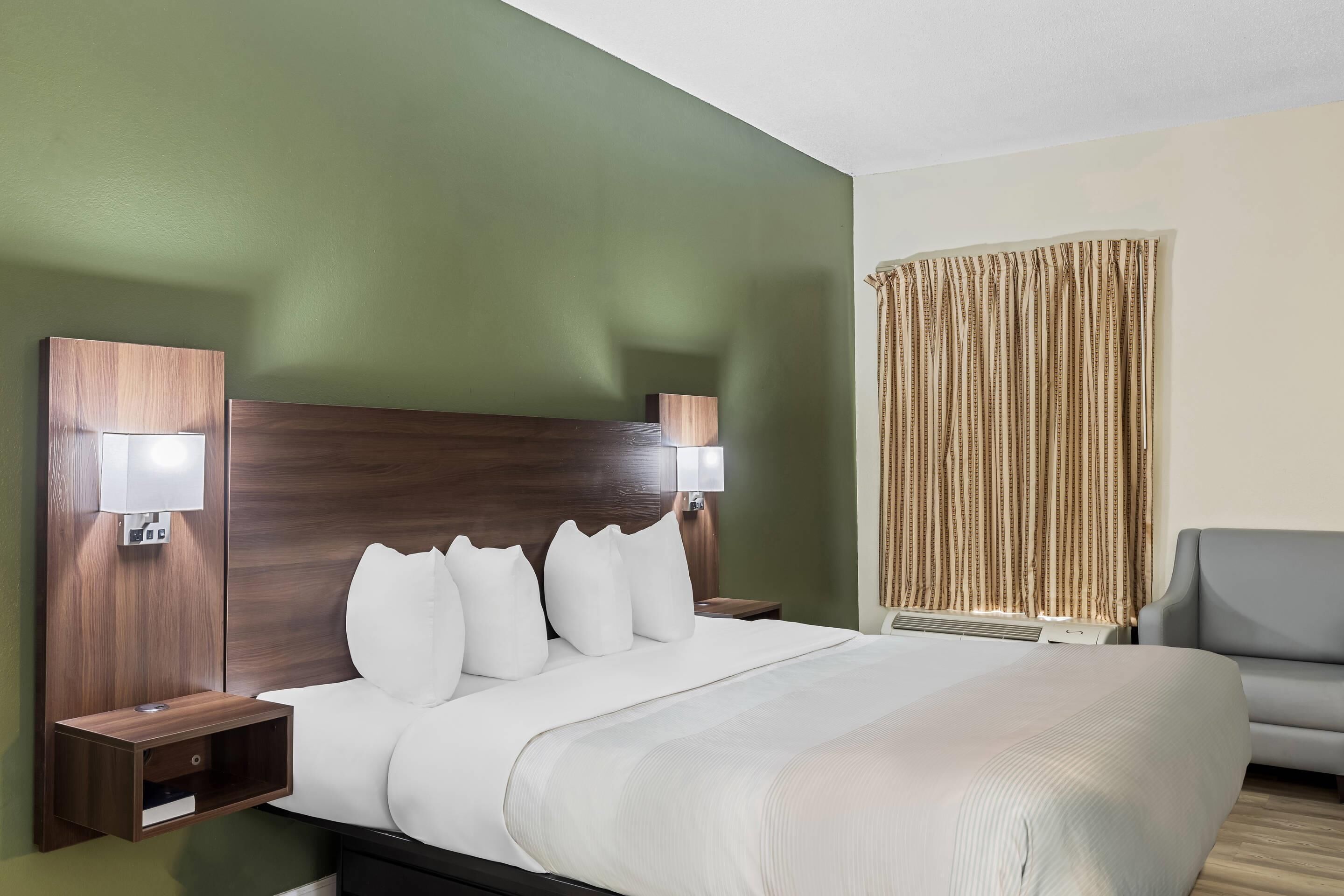A hotel room with a large bed, two bedside tables with lamps, a grey armchair, and a window with curtains.