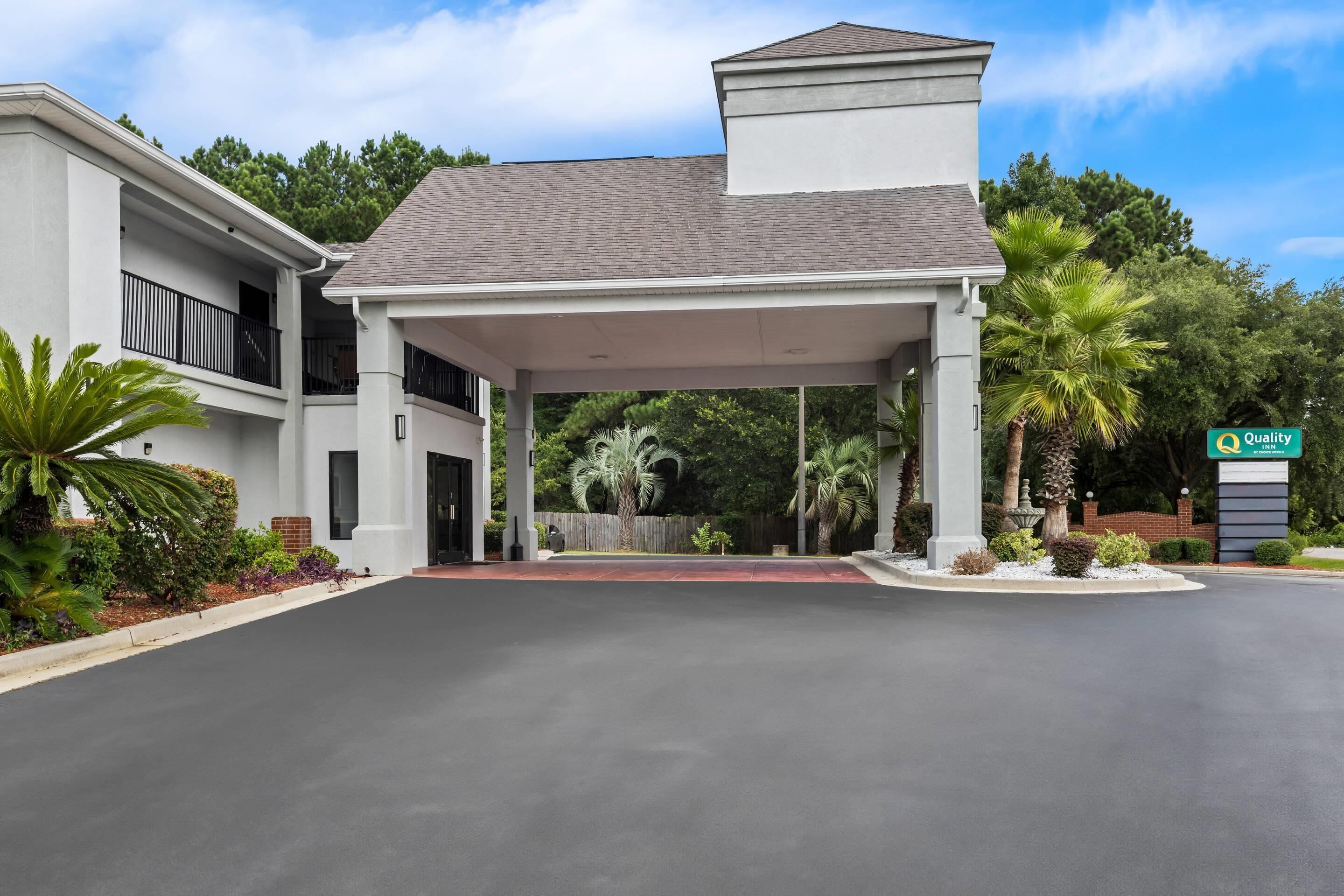 A modern hotel entrance with a covered walkway, palm trees, and a sign indicating 'Quality'.