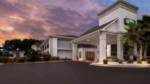 Un hotel con entrada cubierta, un letrero que indica que es un Quality Inn, y una zona ajardinada con palmeras y arbustos.
