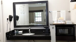 A bathroom with a black countertop, a sink, a mirror, a hairdryer, a towel rack, and a microwave.