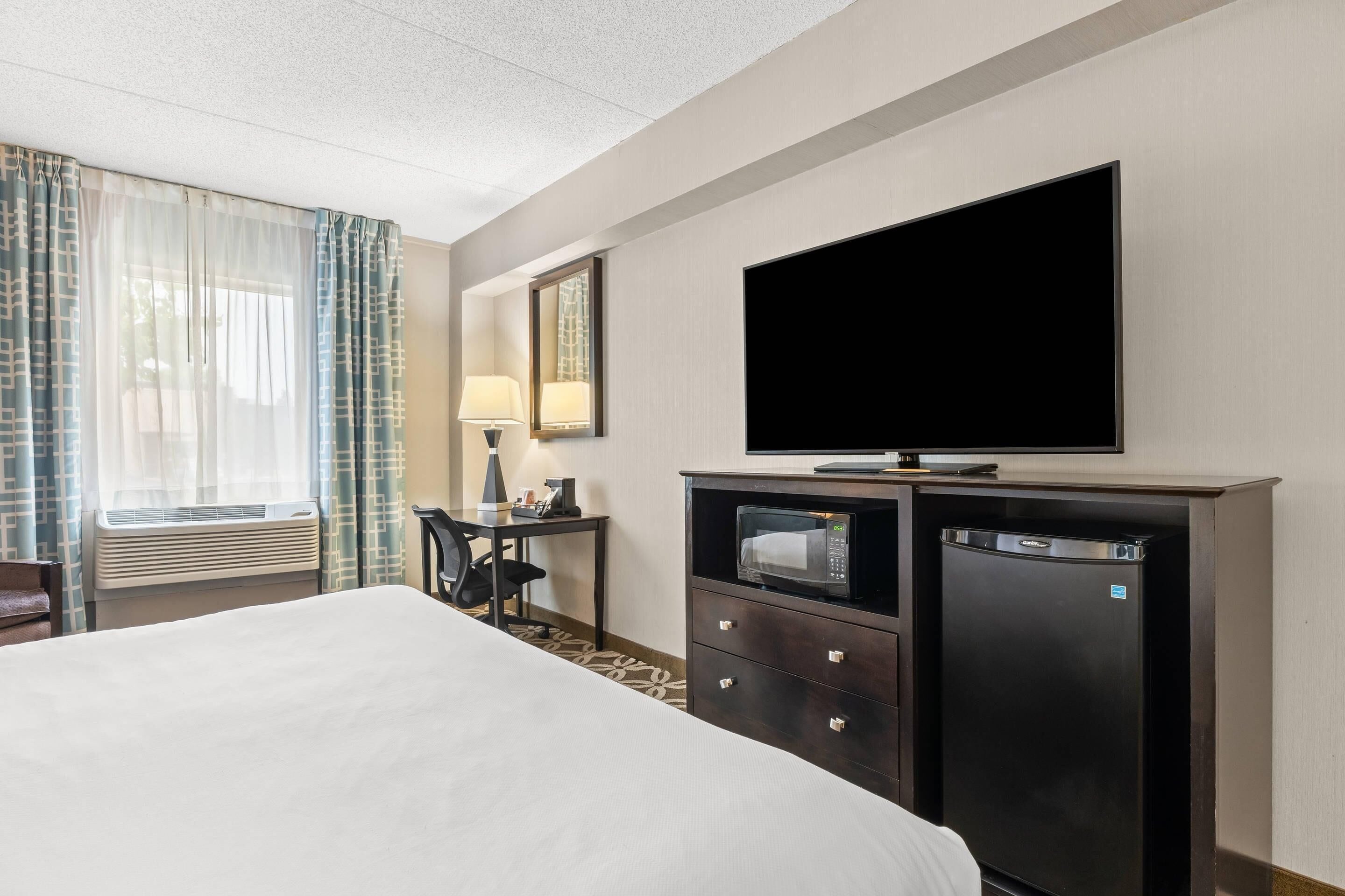 A hotel room with a bed, a desk with a lamp, a television, a microwave, and a refrigerator.