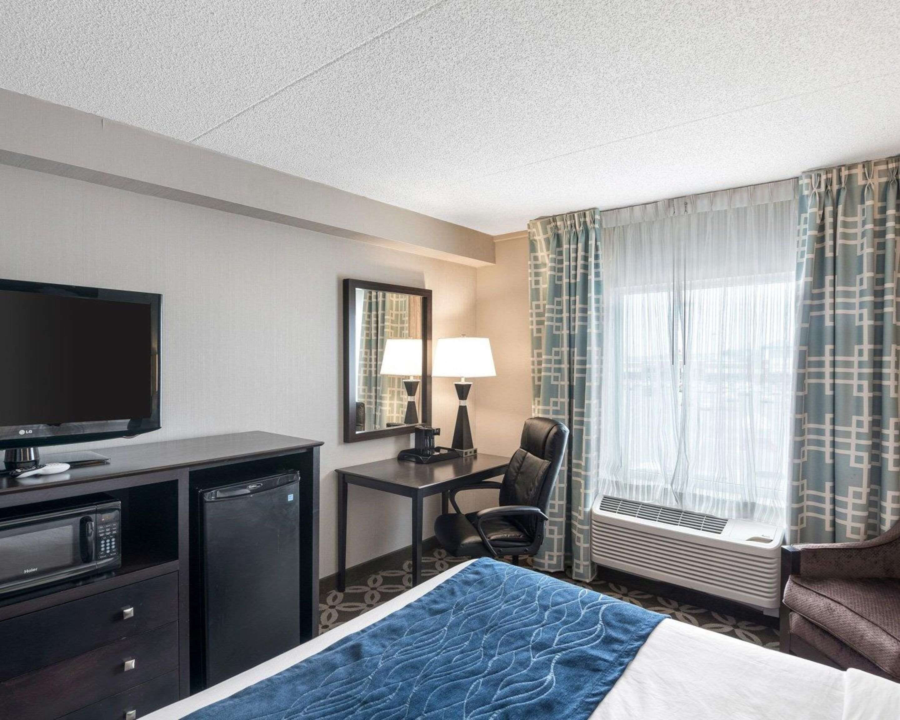 A hotel room with a bed, a desk with a computer, a television, a chair, a mirror, and a window with curtains.