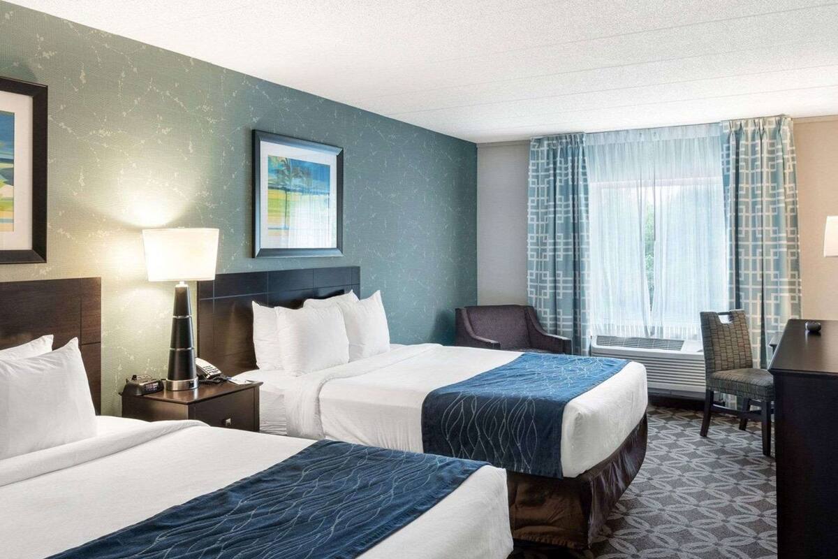 A hotel room with two beds, a desk, a chair, a lamp, a framed picture, and a window with curtains.