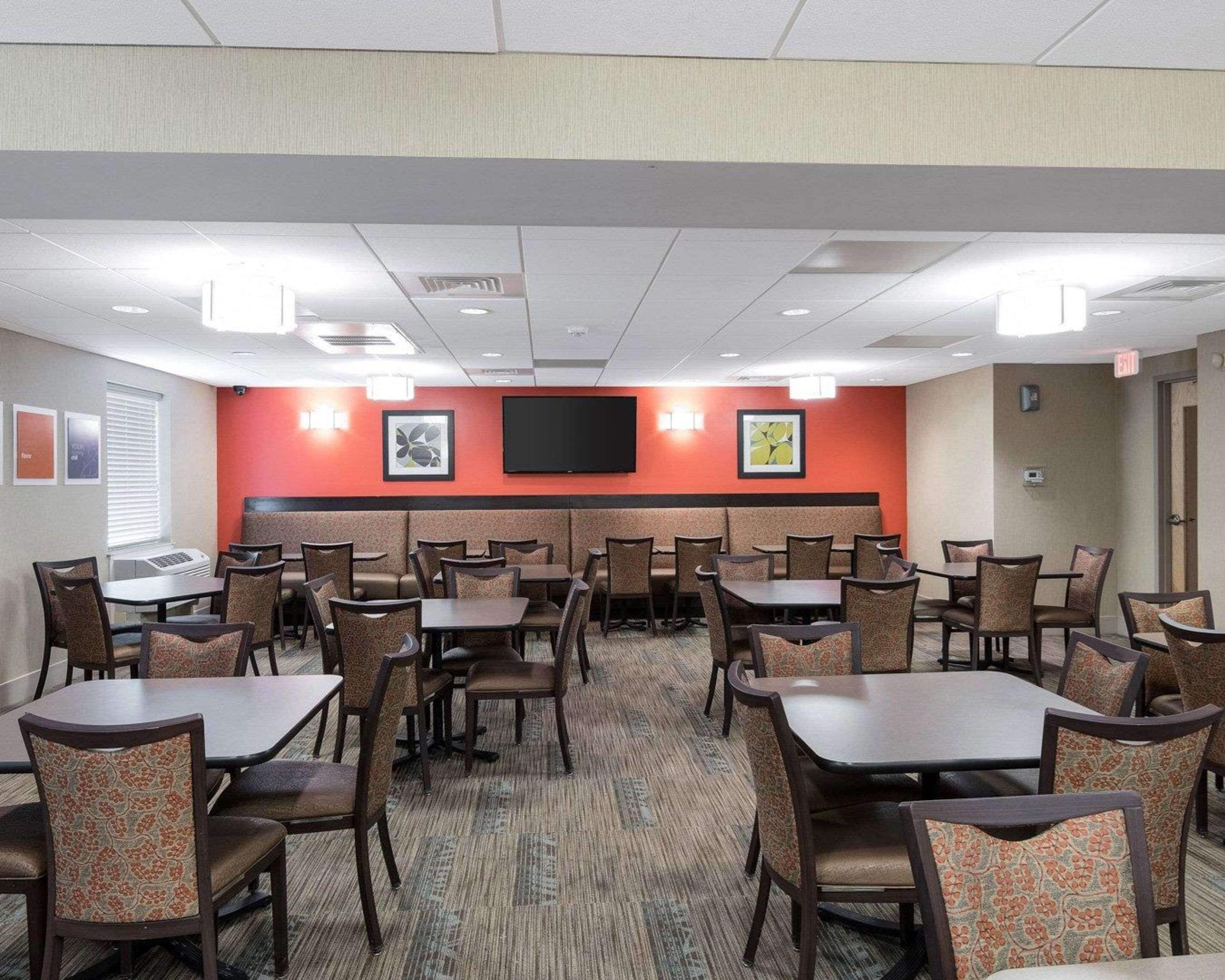 A hotel dining area with multiple tables and booths, a flat-screen TV mounted on the wall, and a carpeted floor.