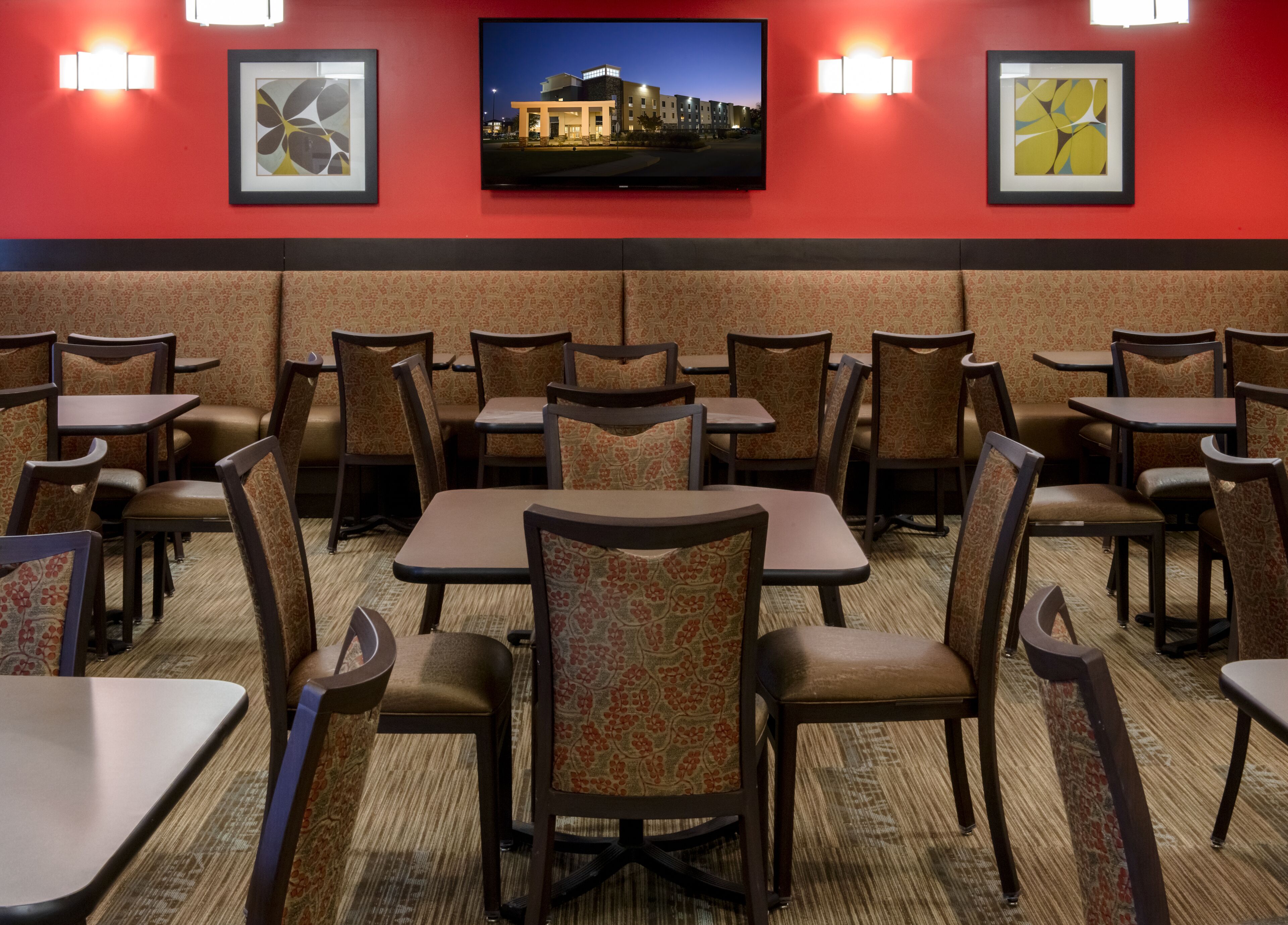 Habitación de hotel con un televisor de pantalla plana grande montado en la pared, varias mesas y sillas dispuestas en una zona de comedor, y cuadros en las paredes.