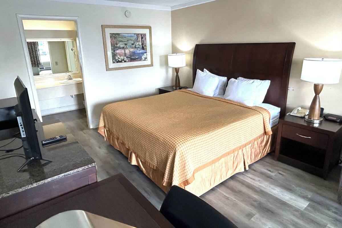 Quarto de hotel com uma cama grande, uma escrivaninha com computador, uma cadeira, mesa de cabeceira com abajur e um quadro na parede.