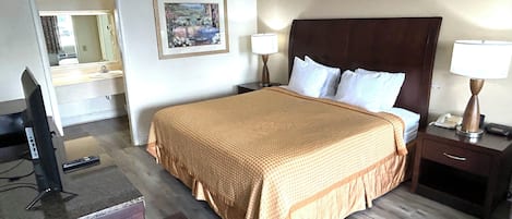Habitación de hotel con una cama grande, un escritorio con una computadora, una silla, una mesita de noche con una lámpara y un cuadro en la pared.
