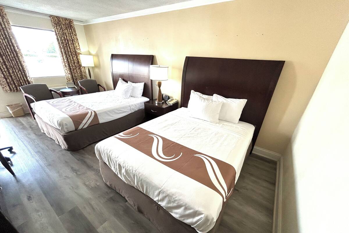 Quarto de hotel com duas camas, uma escrivaninha, uma cadeira e um abajur.