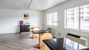 A clean, modern room with a tiled floor, a black marble table, two wooden chairs, a small kitchenette with a fridge and a coffee machine, and a window with a view of a parking lot and buildings.