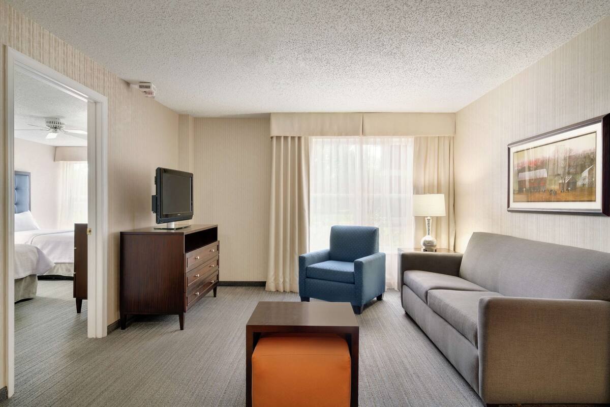 A hotel room with a bed, a television, a blue armchair, a grey sofa, a wooden coffee table, and a framed picture on the wall.