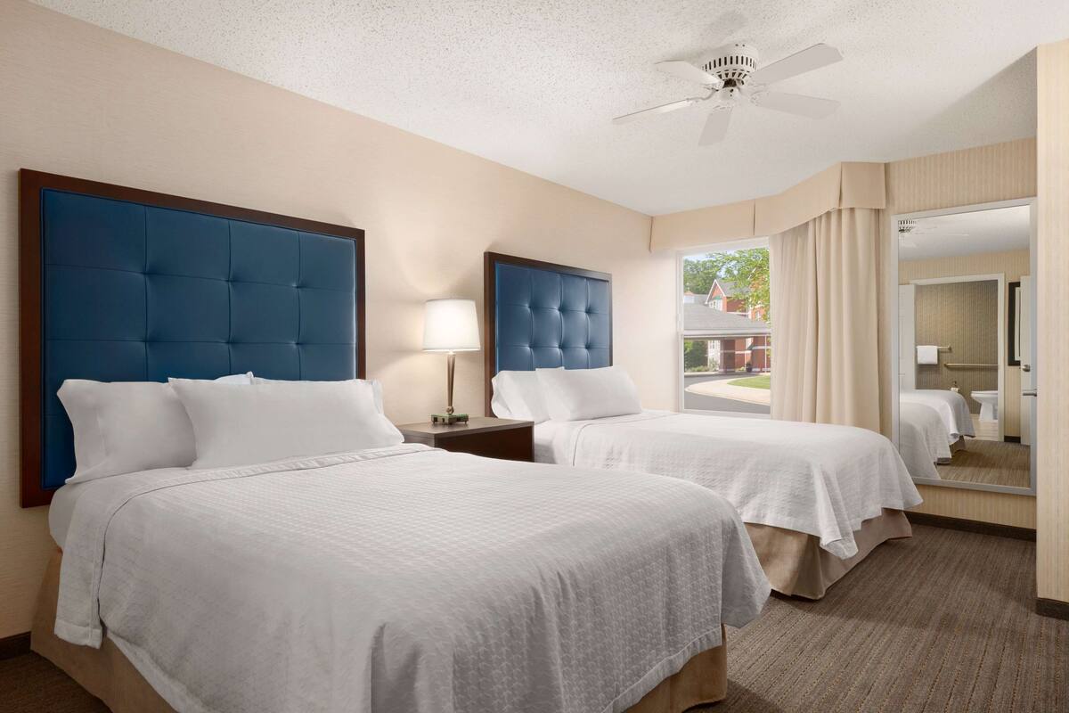 A hotel room with two beds, a ceiling fan, a large mirror, and a view of the outside.