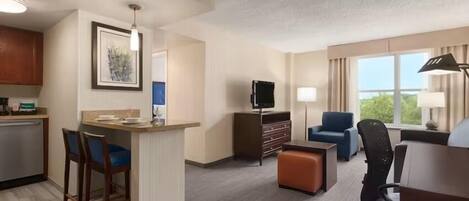 Een hotelkamer met een kitchenette, een televisie, een zithoek met een fauteuil en poef, en een raam met gordijnen.