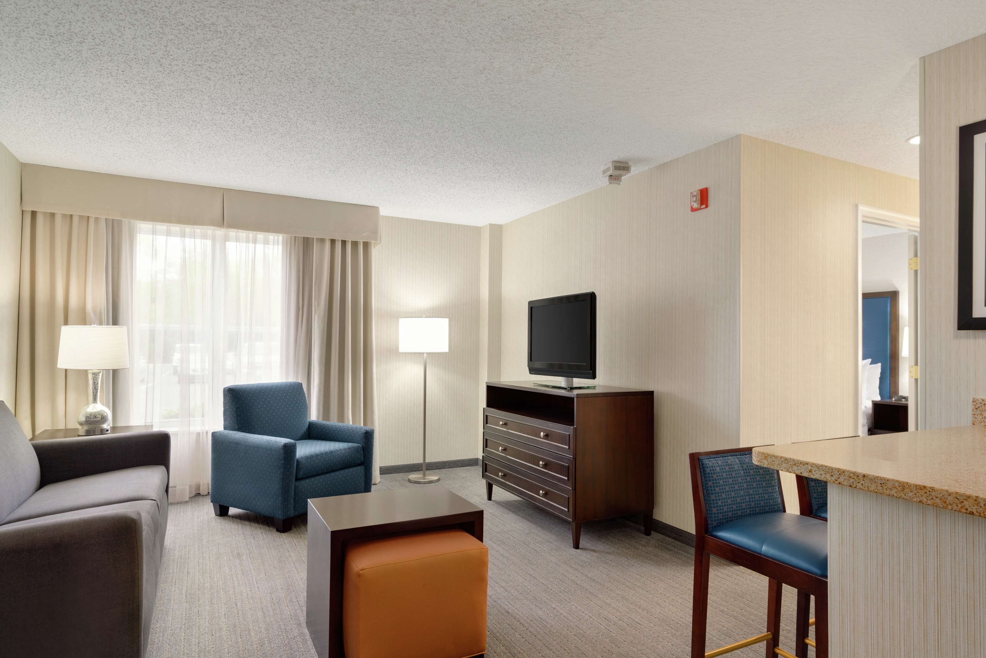 A hotel room with a sofa, armchair, ottoman, TV, dresser, and a small bar area.