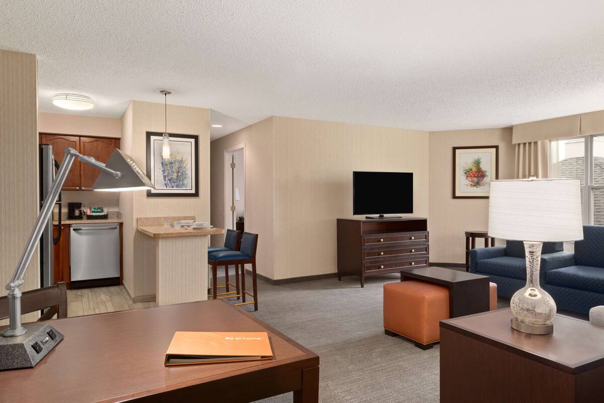A hotel room with a kitchenette, a TV, a sofa, a desk, and a lamp.