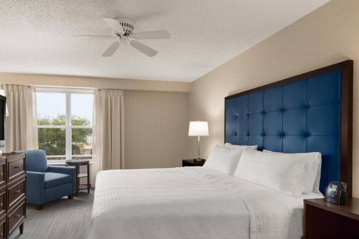 A hotel room with a large bed, a desk, a chair, a ceiling fan, a window, and a lamp.