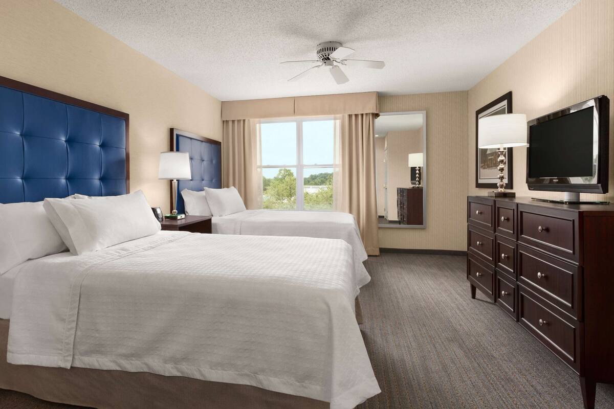 A hotel room with two beds, a television, a dresser, and a large window with curtains.