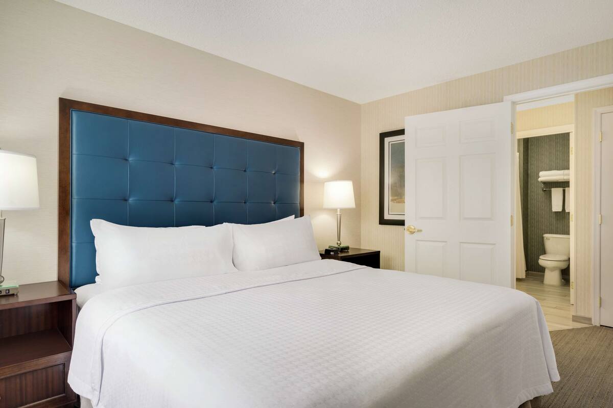 A hotel room with a large bed, a wooden nightstand, a bedside lamp, and a door leading to a bathroom.