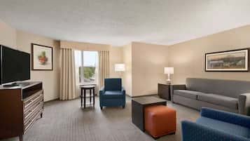 Quarto de hotel com televisão de tela plana, sofá, poltrona azul, banquinho laranja, mesa pequena e luminária.