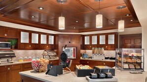 A kitchen with wooden cabinets, a microwave, waffle maker, and a breakfast bar with various food items. - Homewood Suites by Hilton Wilmington-Brandywine Valley (Wilmington)