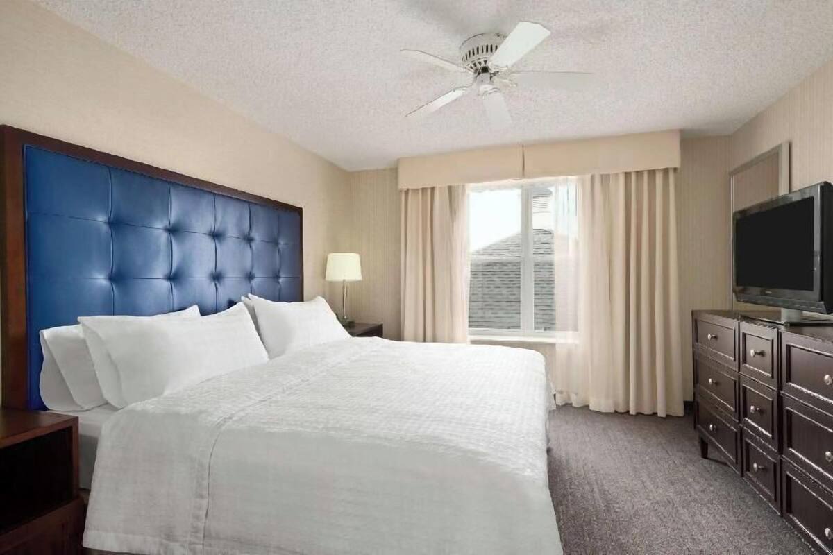 A hotel room with a large bed, a ceiling fan, a TV, and a window with curtains.
