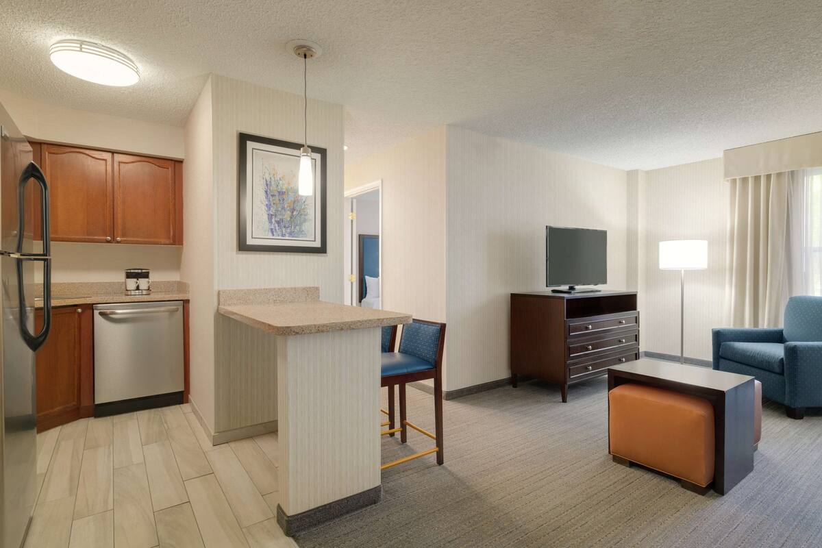 A compact hotel room with a kitchenette, a seating area with a TV, and a window with curtains.