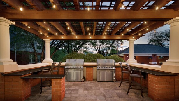 An outdoor patio with two barbecue grills, a pergola with string lights, and a brick wall. - Homewood Suites by Hilton Wilmington-Brandywine Valley (Wilmington)