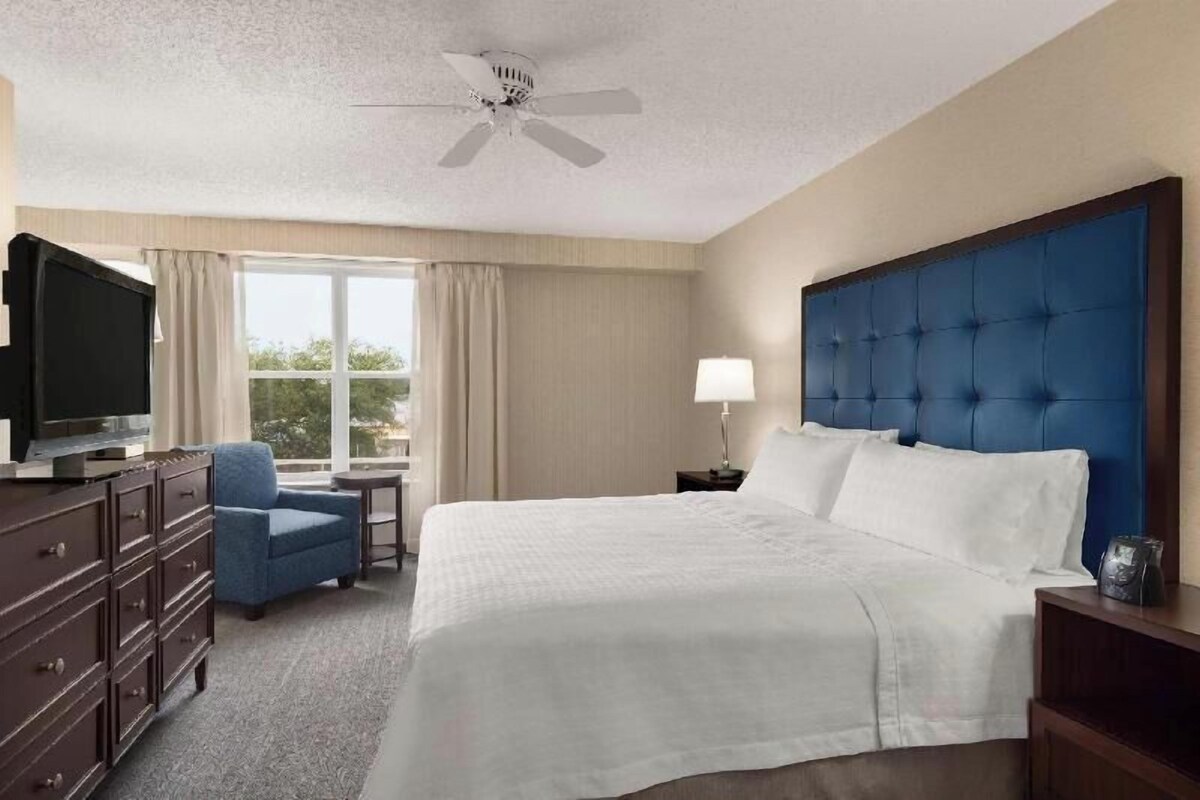 Una habitación de hotel con cama, televisión, escritorio, silla y ventilador de techo.