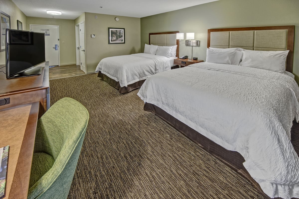 A hotel room with two beds, a desk with a flat-screen TV, a green chair, and a carpeted floor.
