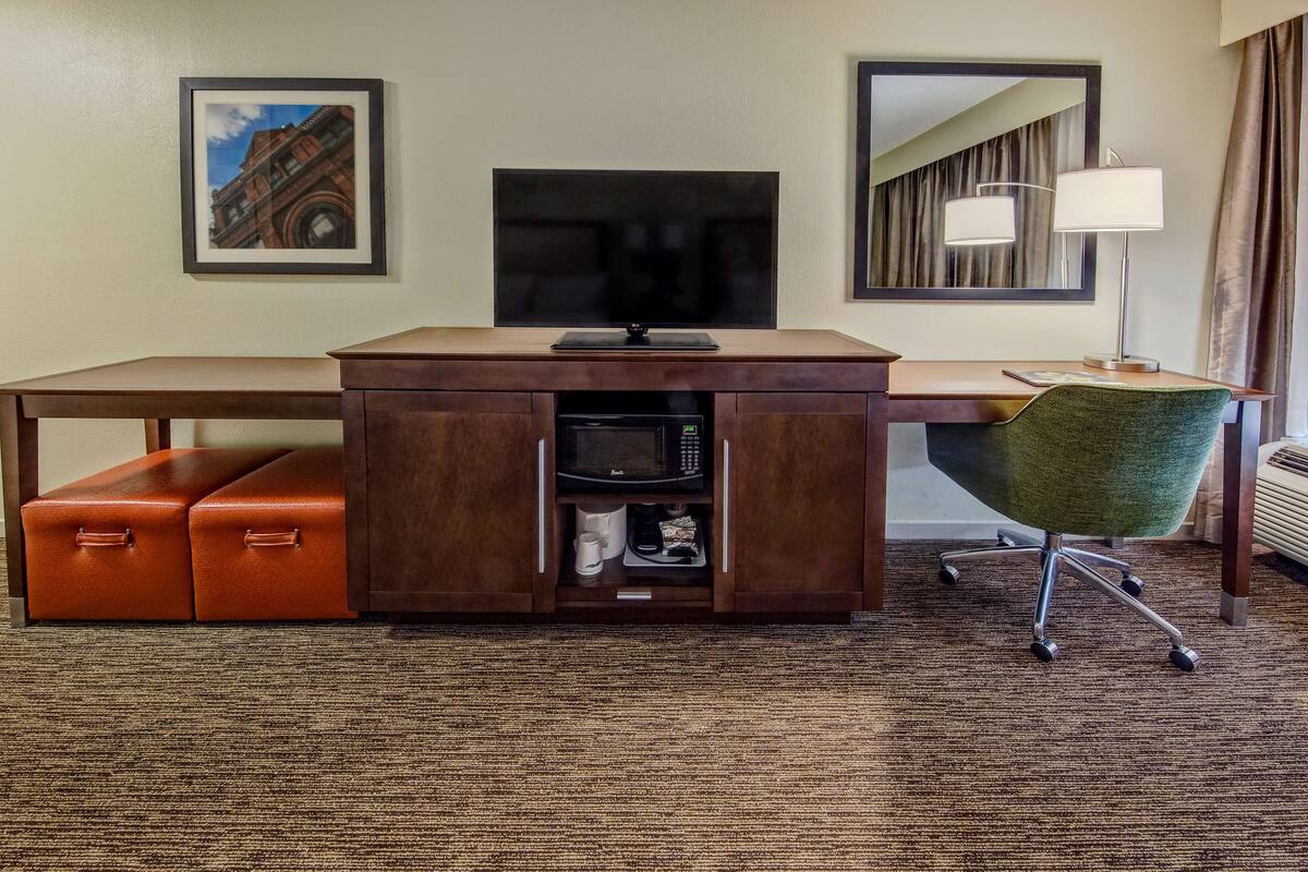 A hotel room with a wooden desk, a green chair, a flat-screen TV, a framed picture, a lamp, and a microwave.