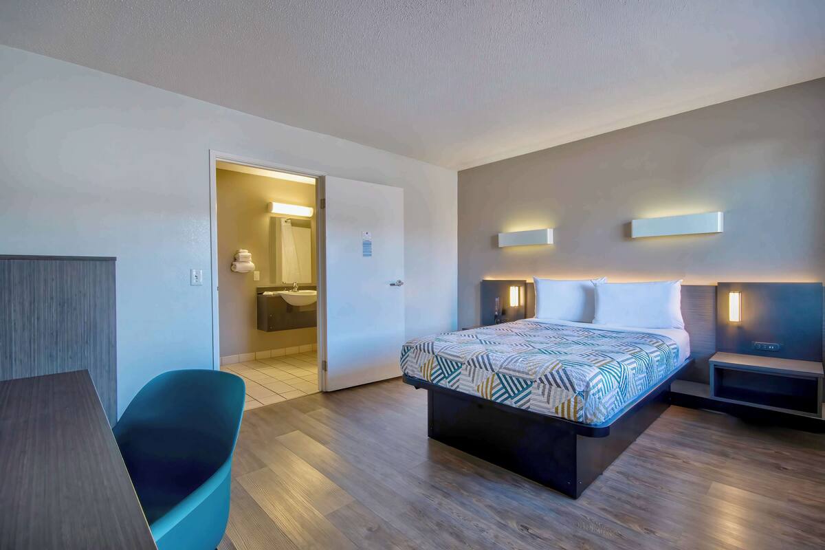 Une chambre d’hôtel moderne, dotée d’un grand lit, d’un bureau et d’une chaise. On peut apercevoir la salle de bain à travers une porte ouverte.