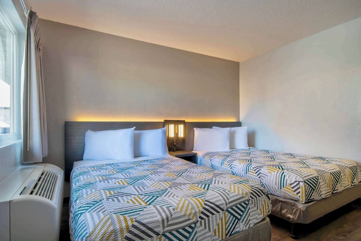 Une chambre d’hôtel avec deux lits, une table de chevet, une lampe et une fenêtre avec des rideaux.