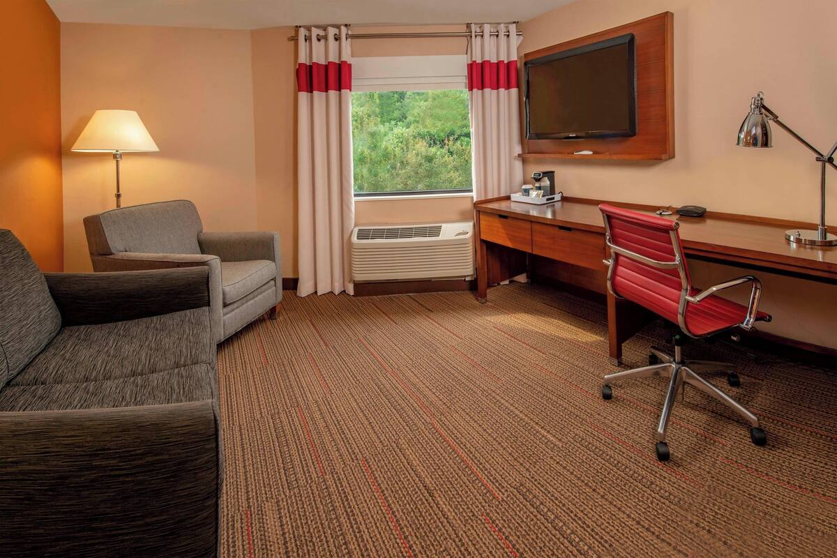 A hotel room with a sofa, a desk, a chair, a TV, and an air conditioning unit.