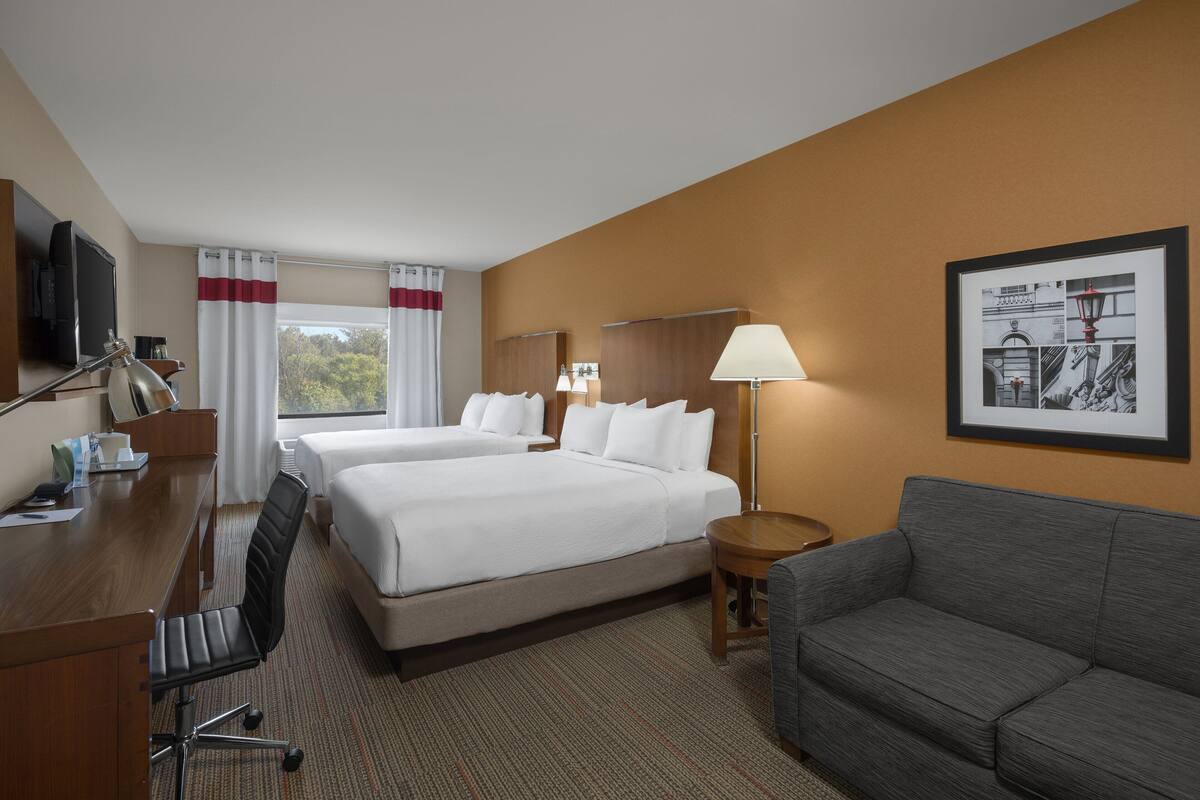 A hotel room with two beds, a desk, a chair, a sofa, a lamp, and a framed picture on the wall.