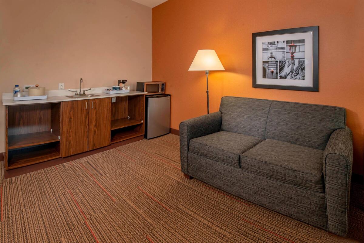 A hotel room with a kitchenette, a grey sofa, a lamp, and a framed picture on the wall.