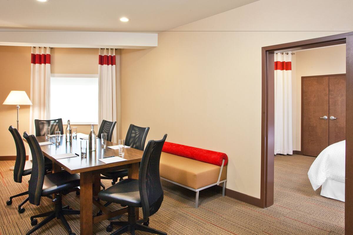 A hotel room with a conference table, chairs, a sofa, a bed, and a door leading to another room.