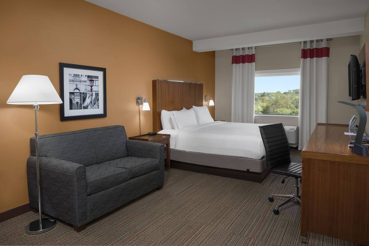 A hotel room with a bed, a desk, a chair, a sofa, a lamp, and a framed picture on the wall.