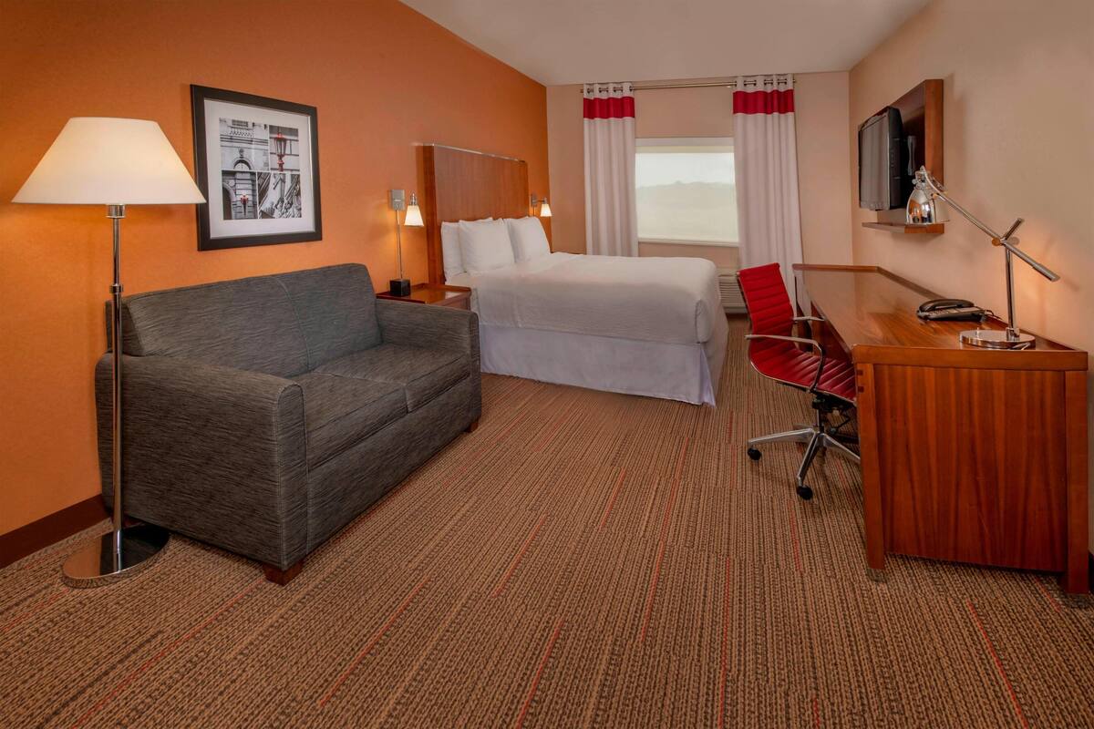 A hotel room with a bed, a desk, a chair, a lamp, and a picture on the wall.