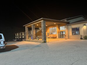 Un edificio de dos pisos con una zona de estar al aire libre cubierta, pilares de piedra y un estacionamiento, de noche.