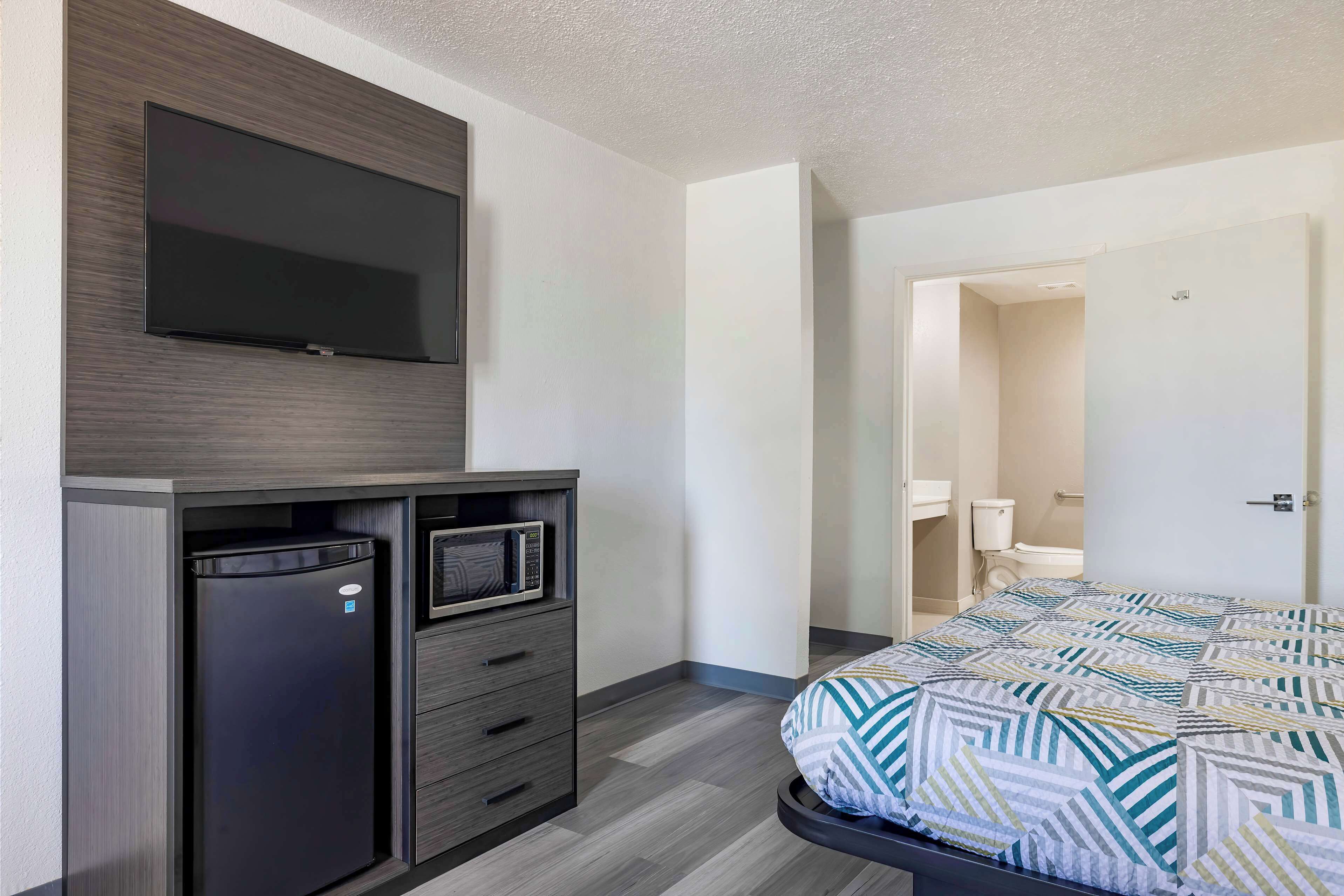 A hotel room with a bed, a TV mounted on the wall, a refrigerator, and a microwave.