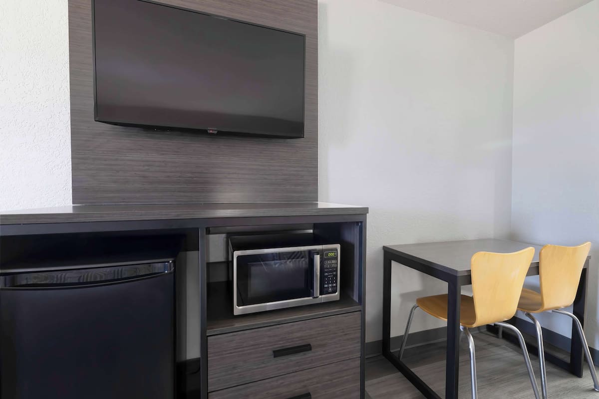 A compact kitchenette with a microwave, a small refrigerator, and a flat-screen TV mounted on the wall. There are two chairs and a small table.