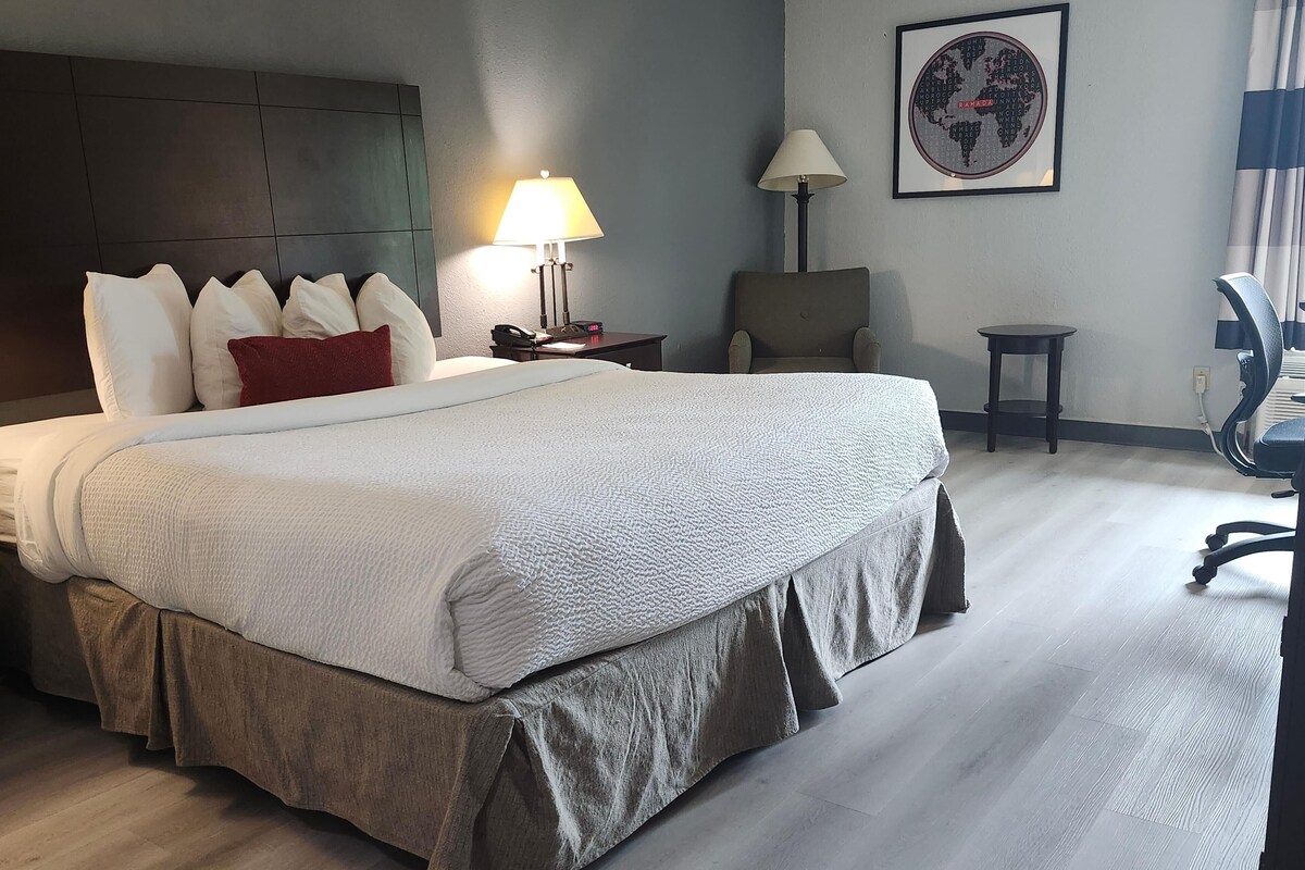 A hotel room with a large bed, a desk with a lamp, a chair, a small table, and a world map on the wall.